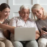 a senior husband and wife sitting on a couch with their adult daughter who is holding a phone all looking at senior living communities on a laptop