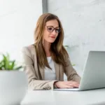 a woman Marketing manager wearing glasses working on her laptop at her desk with a plant on the desk